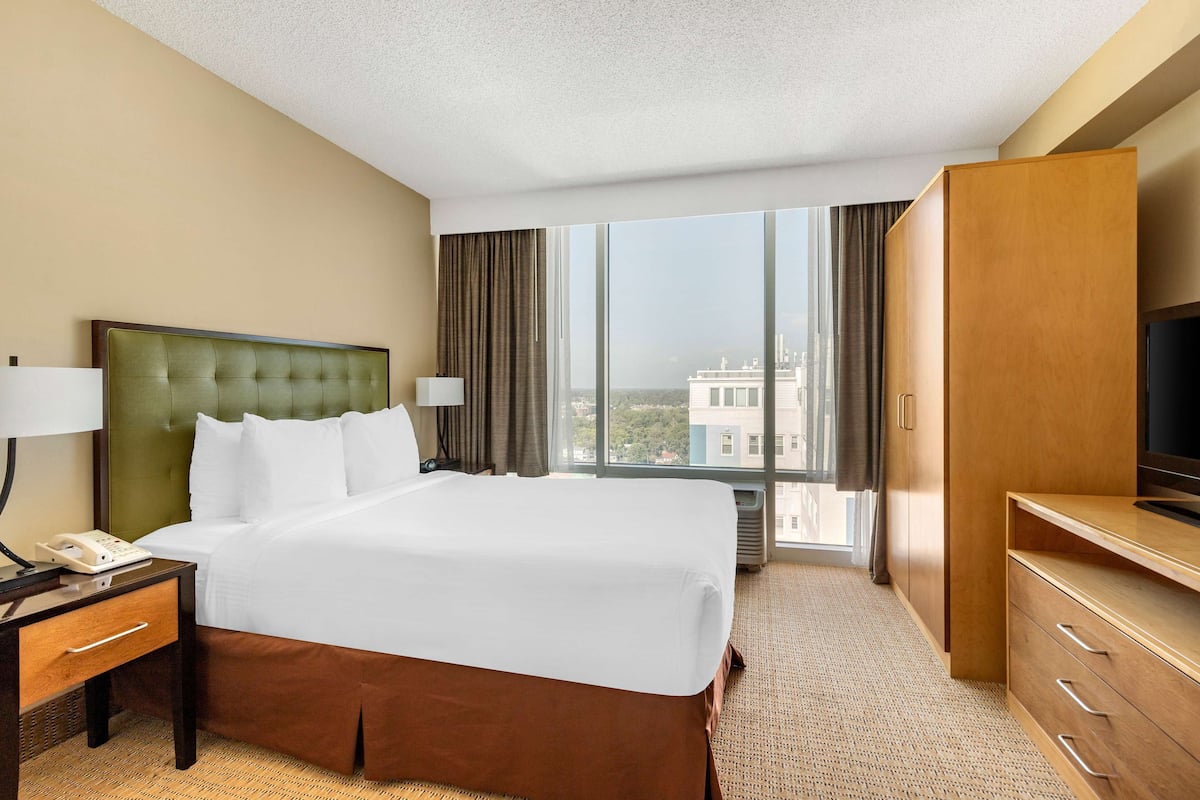 A hotel room with a large bed, a TV, a desk with a phone, and a view of the city through large windows.