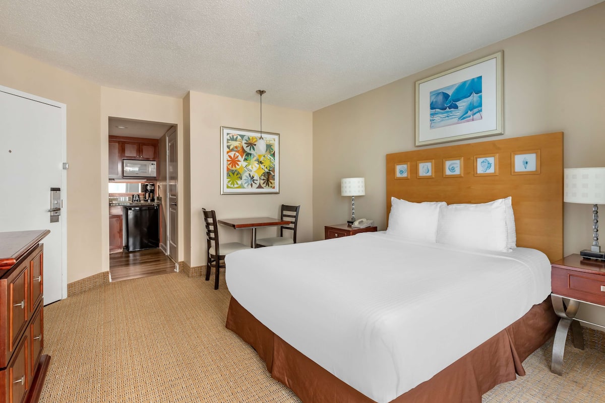 A hotel room with a large bed, a desk, a chair, a small kitchen area, and a dining table.