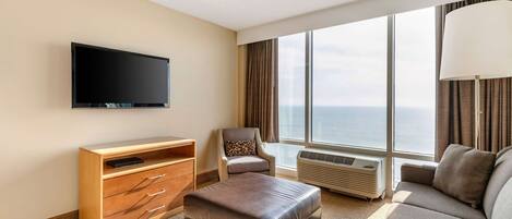 A hotel room with a flat-screen TV, a sofa, a chair, a wooden cabinet, and a large window with a view of the ocean.