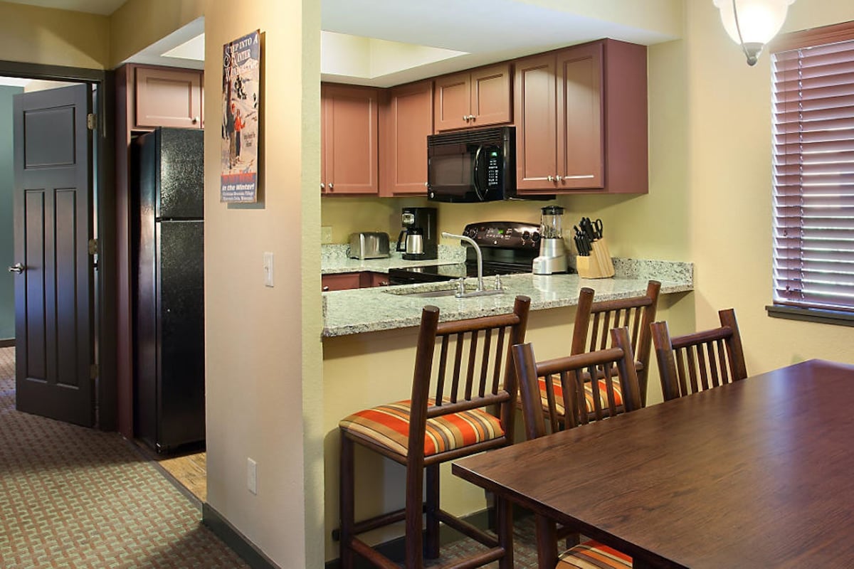 A compact kitchen with wooden cabinets, a microwave, a toaster, and a blender. There is a dining table with chairs and a window with blinds.