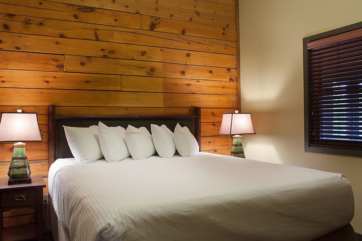 A neatly made bed with white linens and pillows, flanked by two bedside tables with lamps, in a room with wooden paneling on the walls.