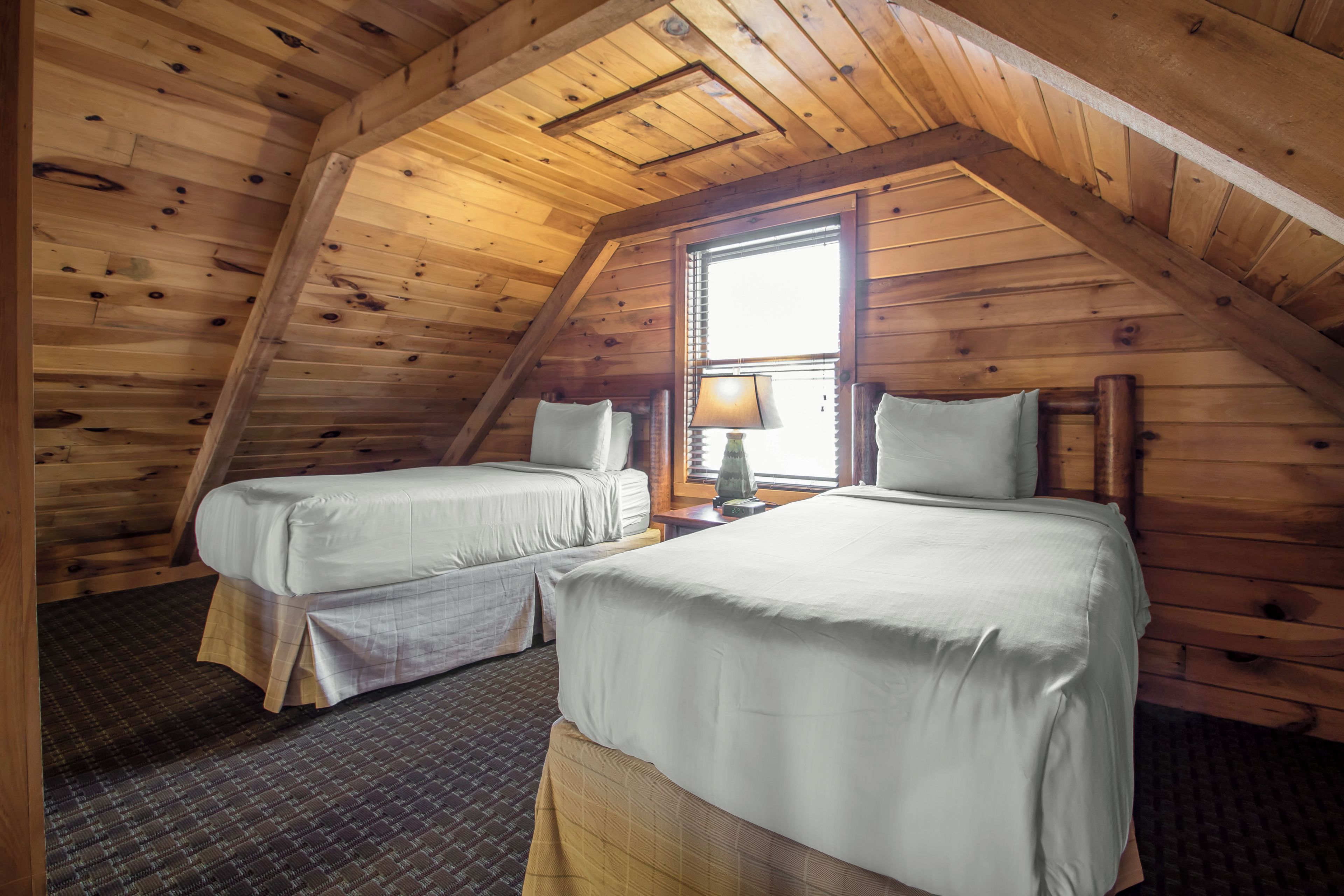 A room with two beds, wooden walls, a window with blinds, and a lamp.