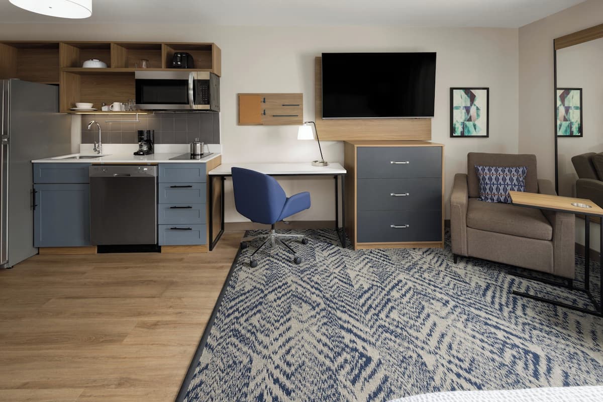 Ein kompaktes Hotelzimmer mit Küchenzeile, einem Flachbildfernseher, einem Schreibtisch, einem Sessel, einer Couch und einem gemusterten Teppich.