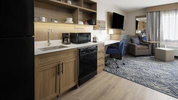 A modern kitchen with a microwave, coffee maker, and dishwasher. There is a seating area with a sofa and armchair, and a flat-screen TV mounted on the wall.
