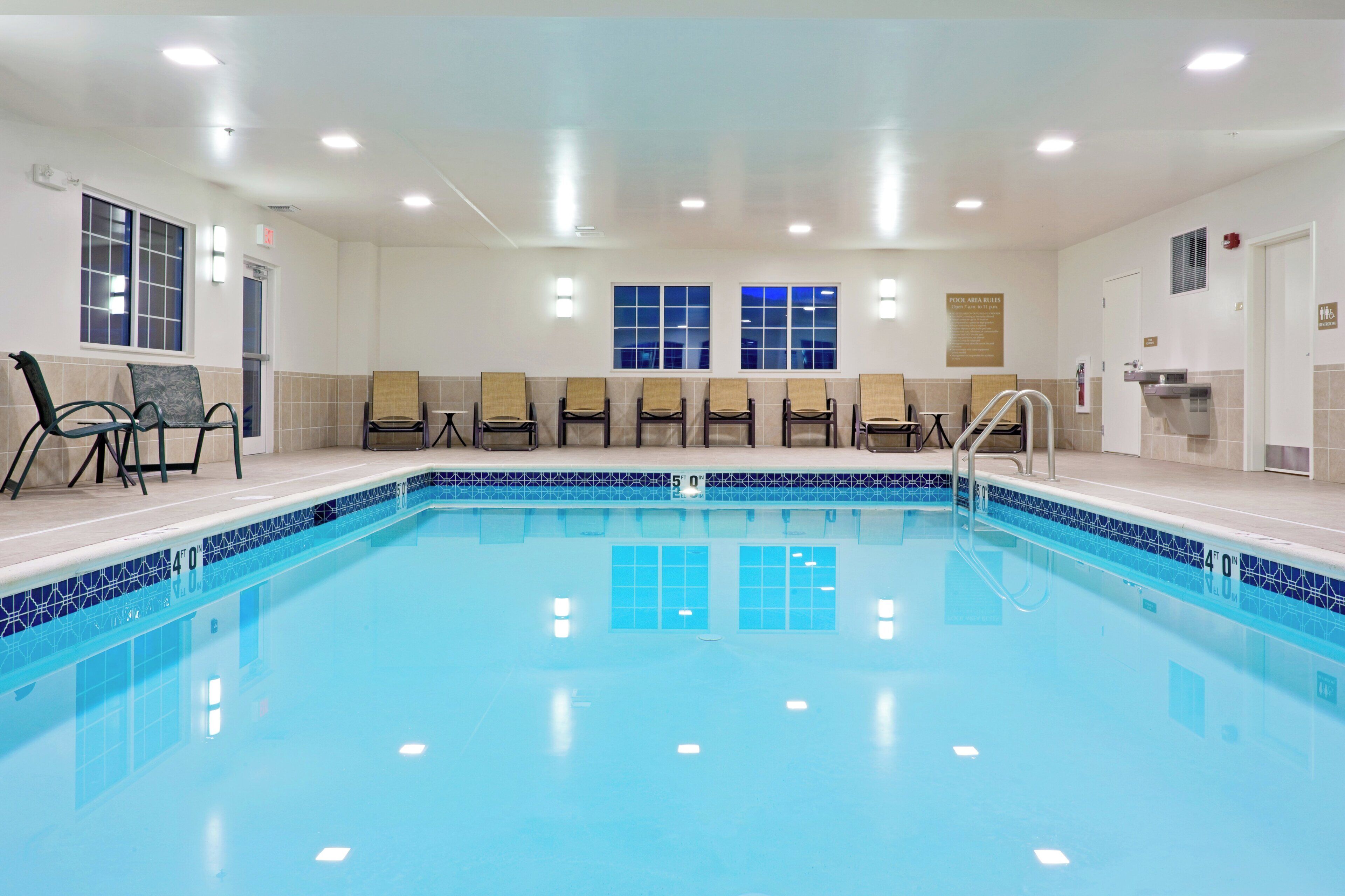 An indoor swimming pool with a tiled edge, a seating area with chairs, and a sign indicating pool area rules.