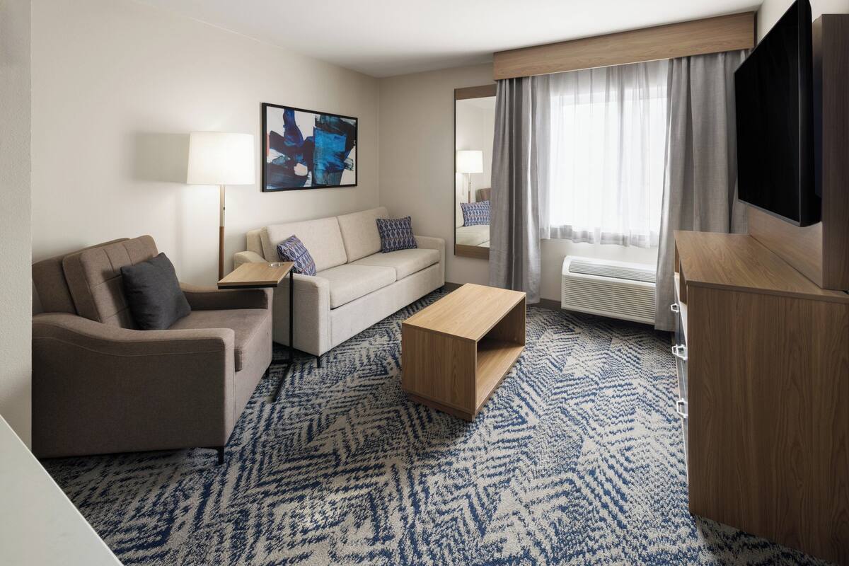A hotel room with a sofa, a coffee table, a TV, a lamp, a painting, a window with curtains, and a patterned carpet.
