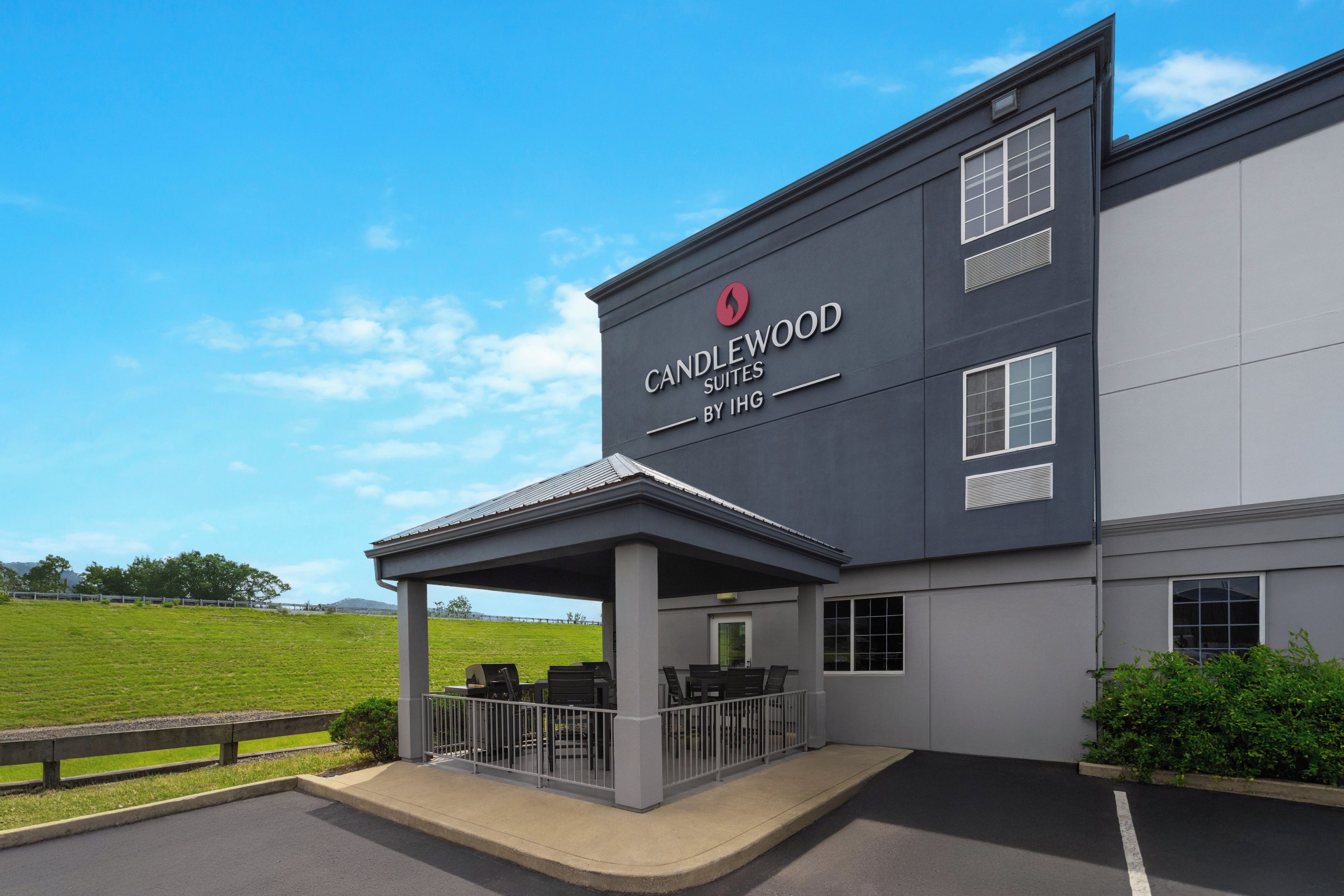 En hotellfasad med ett takförsett utomhusområde med sittplatser, en skylt med texten ”CANDLEWOOD SUITES BY IHG” och en utsikt över en grön äng i bakgrunden.