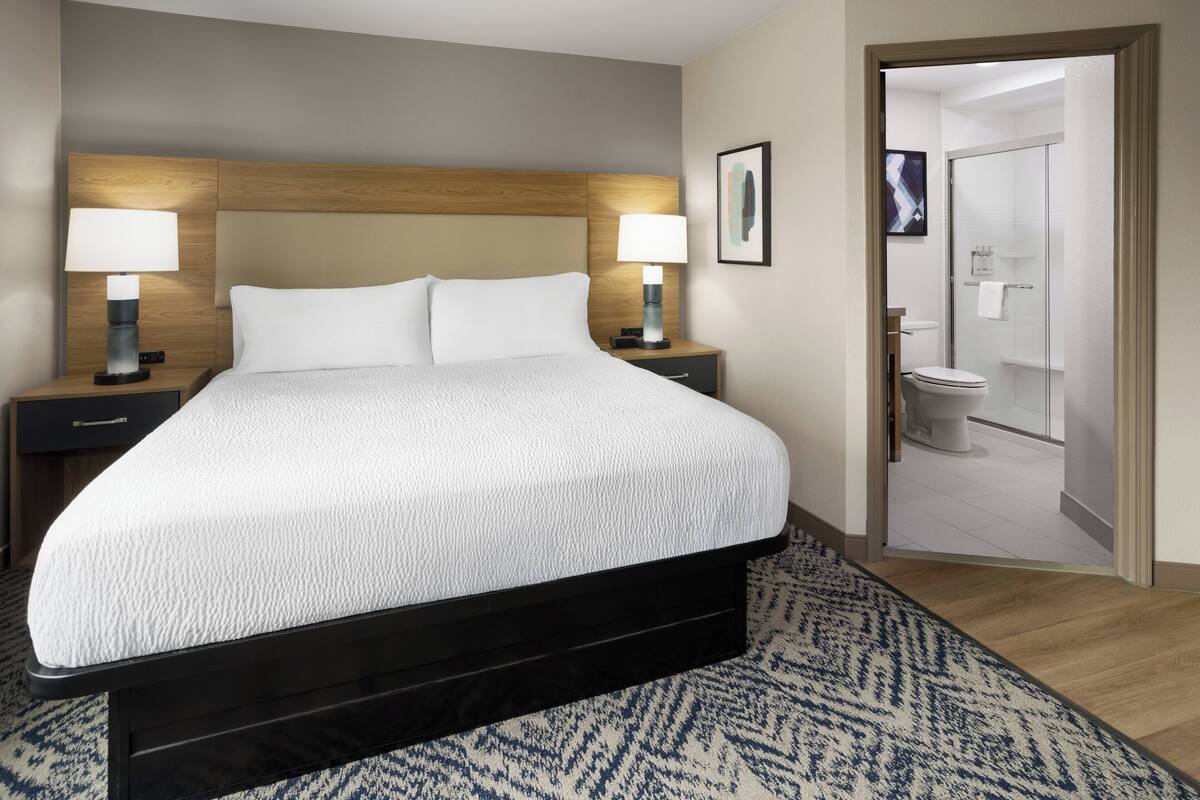 A hotel room with a bed, nightstands, lamps, a bathroom visible through an open door, and a patterned rug.