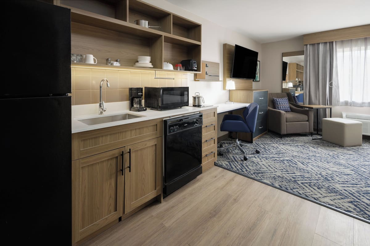 A modern kitchen with a microwave, coffee maker, and dishwasher. There is a seating area with a sofa and armchair, and a flat-screen TV mounted on the wall.