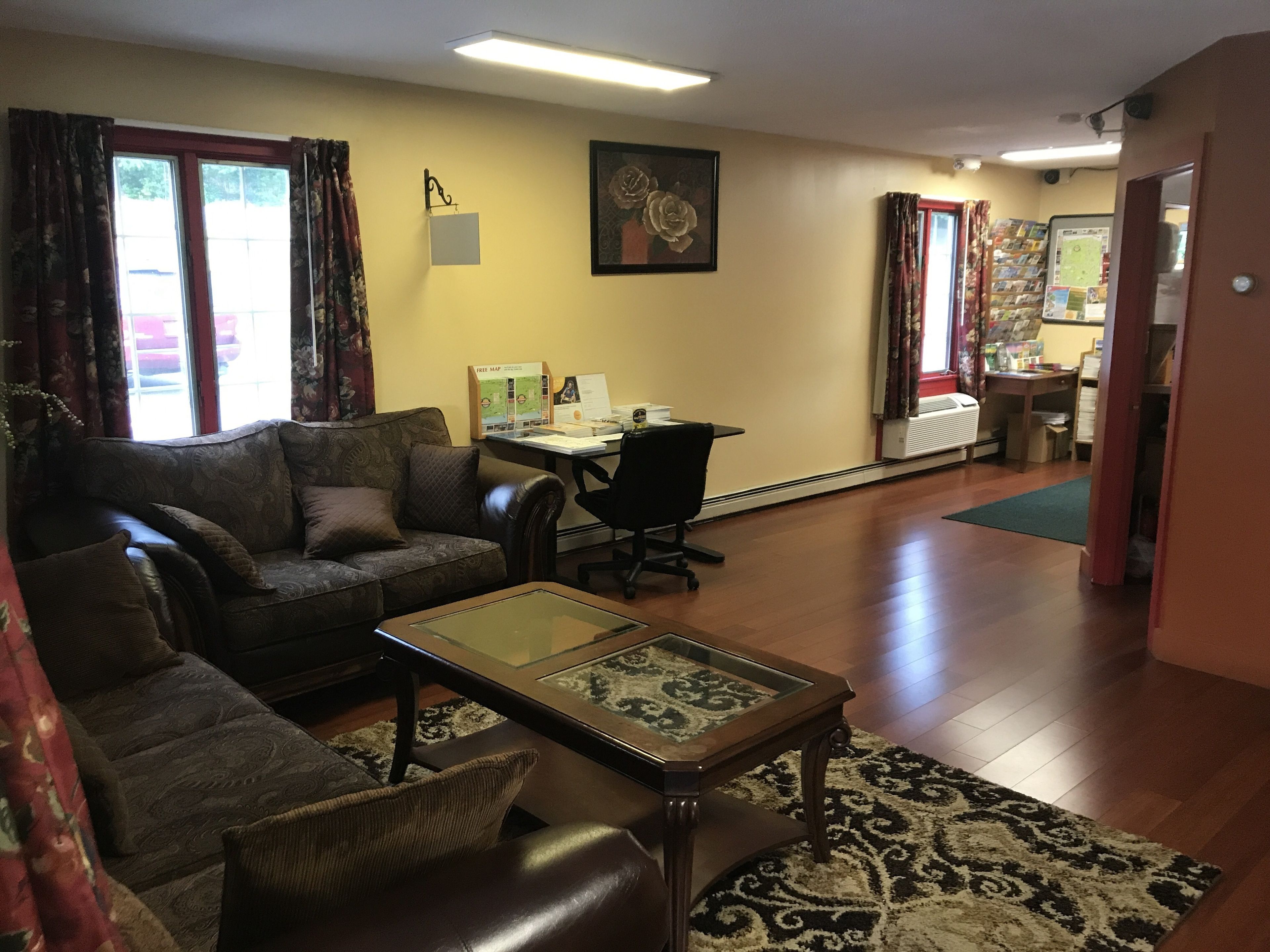 A living room with a sofa, a glass-top coffee table, and a desk with a computer. There is a window with curtains and a radiator.