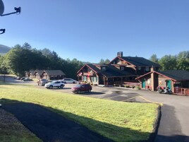 Una cabaña con un amplio porche, varios vehículos estacionados al frente y montañas al fondo.