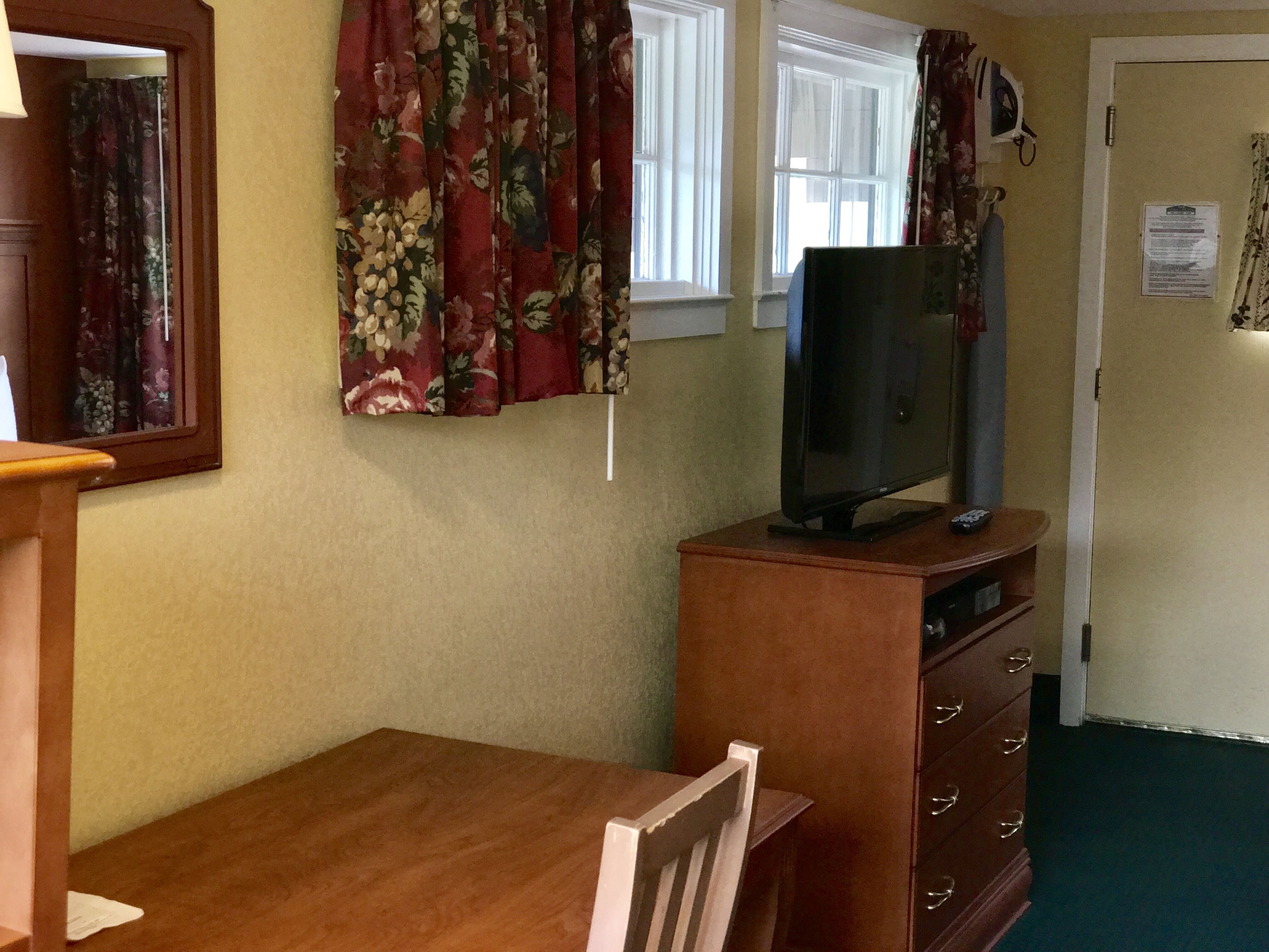 A room with a wooden table, a television on a wooden stand, a mirror, and a window with floral curtains.