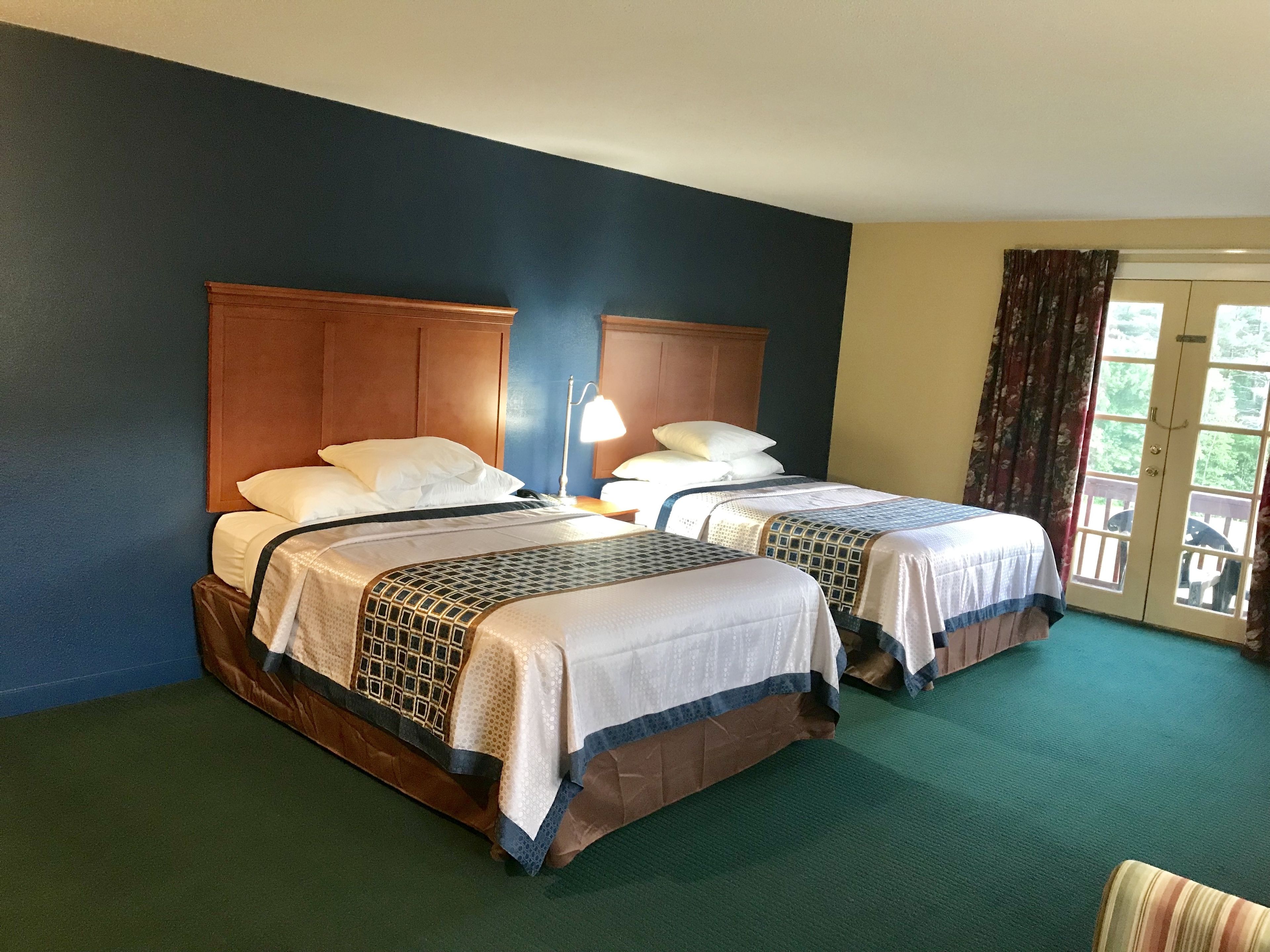 A hotel room with two beds, a wooden headboard, a green carpet, and a view through a door.