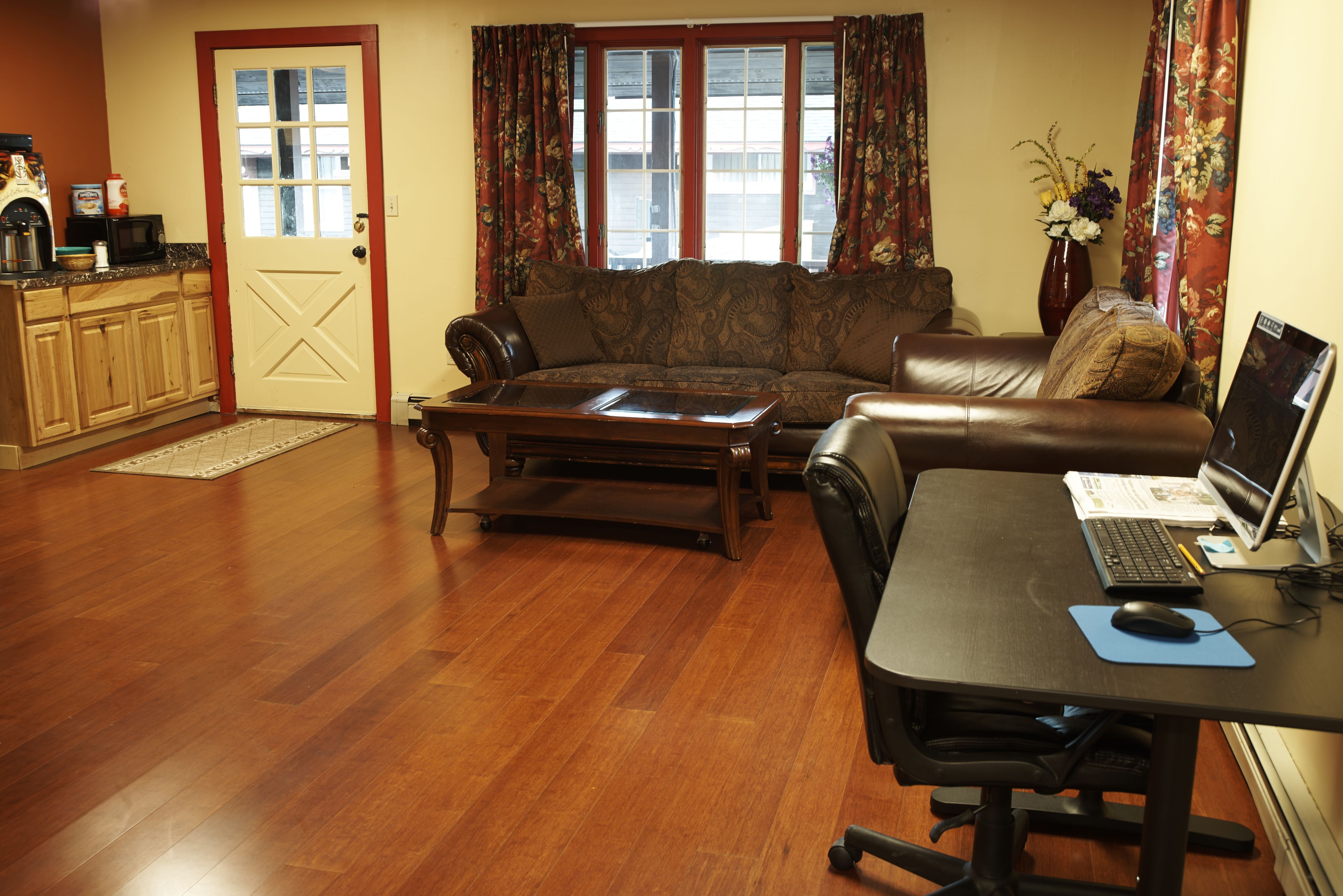 A living room with a wooden floor, a sofa, a coffee table, a desk with a computer, and a window with curtains.