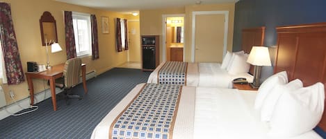 A hotel room with two beds, a desk, a chair, a lamp, and a small kitchenette.