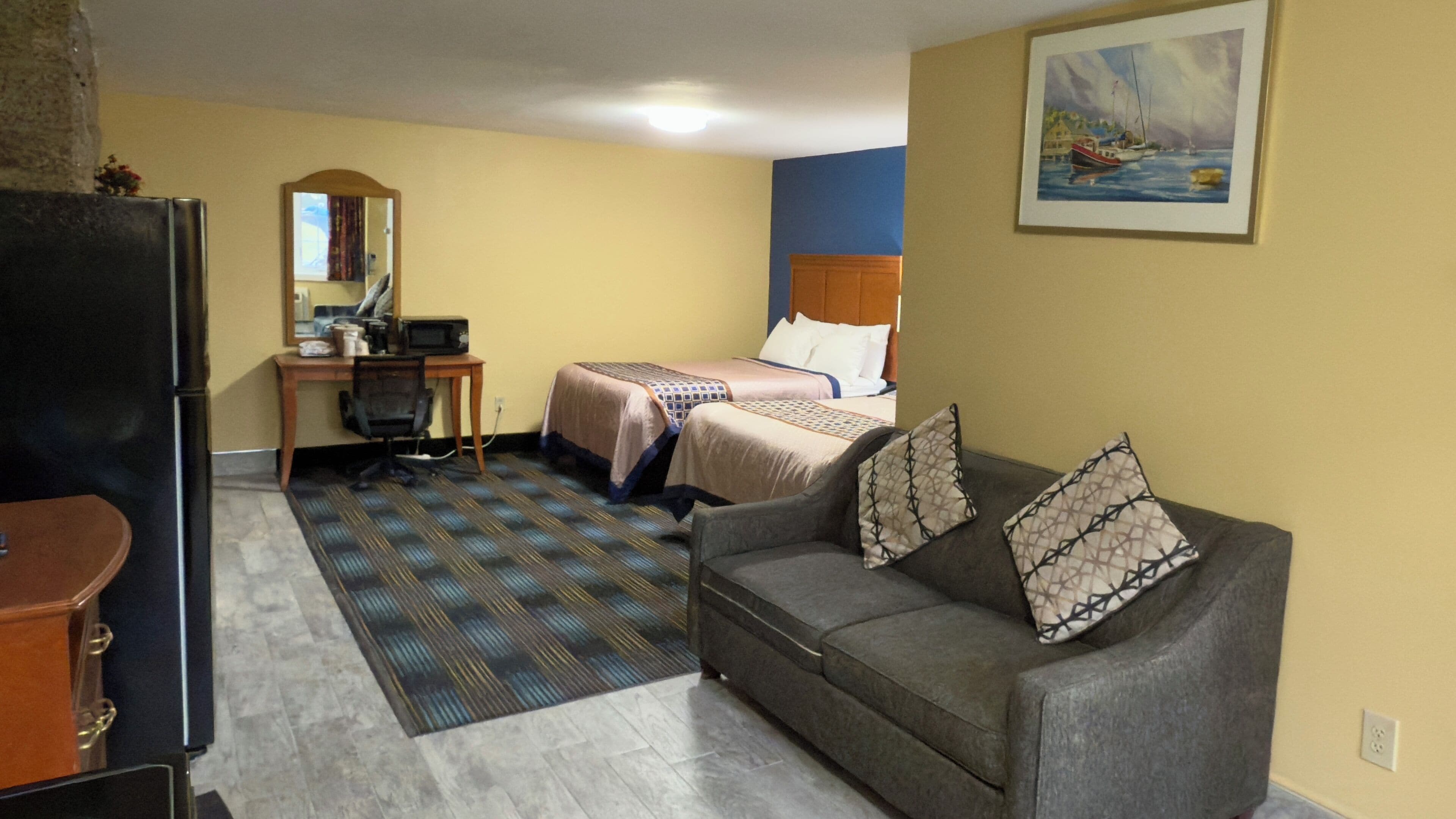 A hotel room with a sofa, two beds, a desk, a refrigerator, and a painting on the wall.