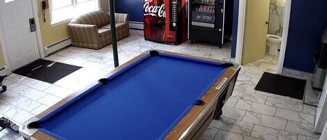 A pool table in a room with a treadmill, a Coca-Cola vending machine, and a door leading to a bathroom.