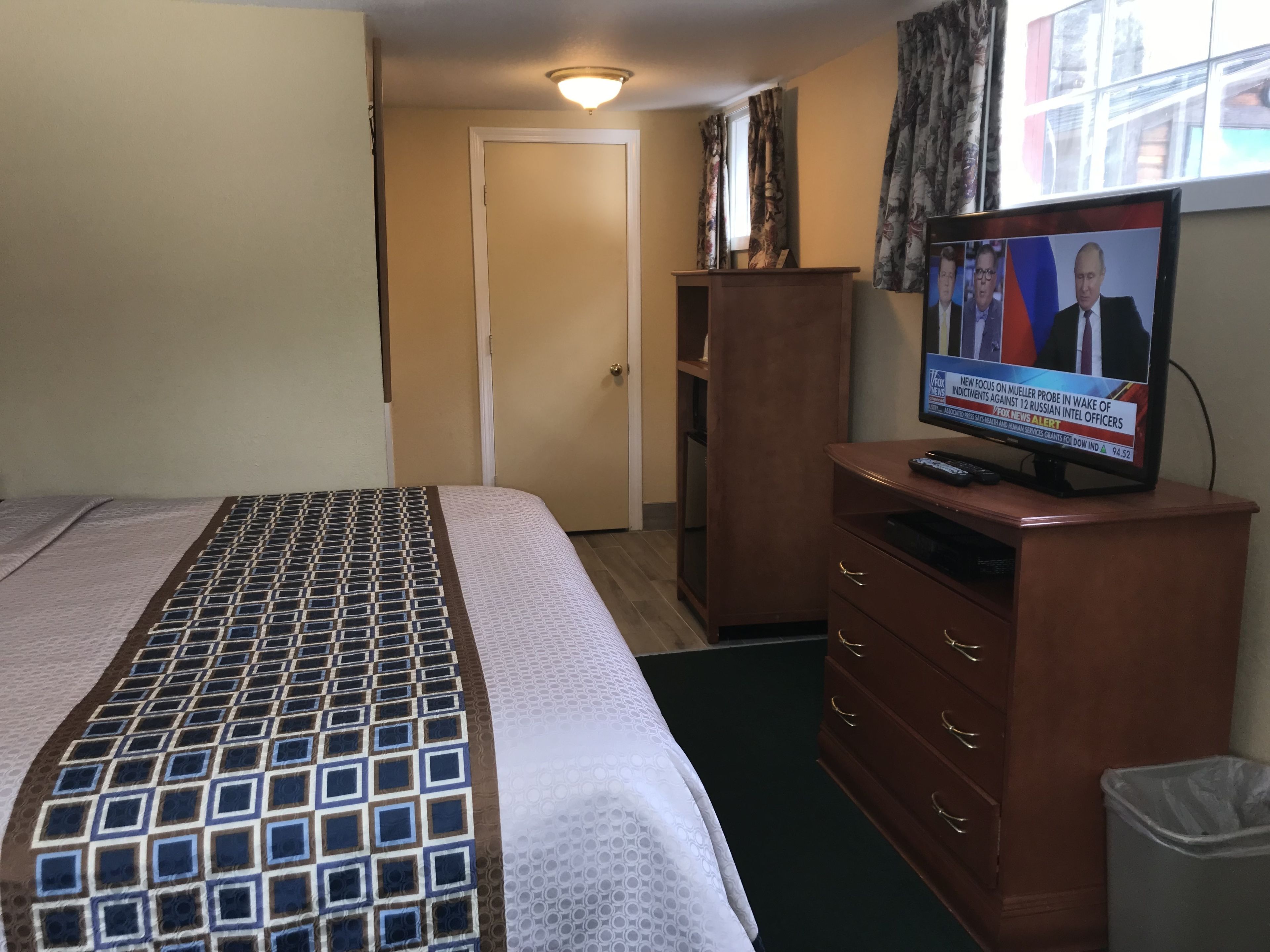 A hotel room with a bed, a wooden dresser, a television displaying a news program, and a door in the background.