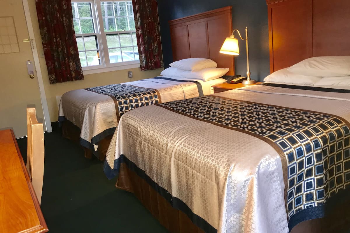 A hotel room with two beds, a nightstand with a lamp, a wooden dresser, and a window with curtains.