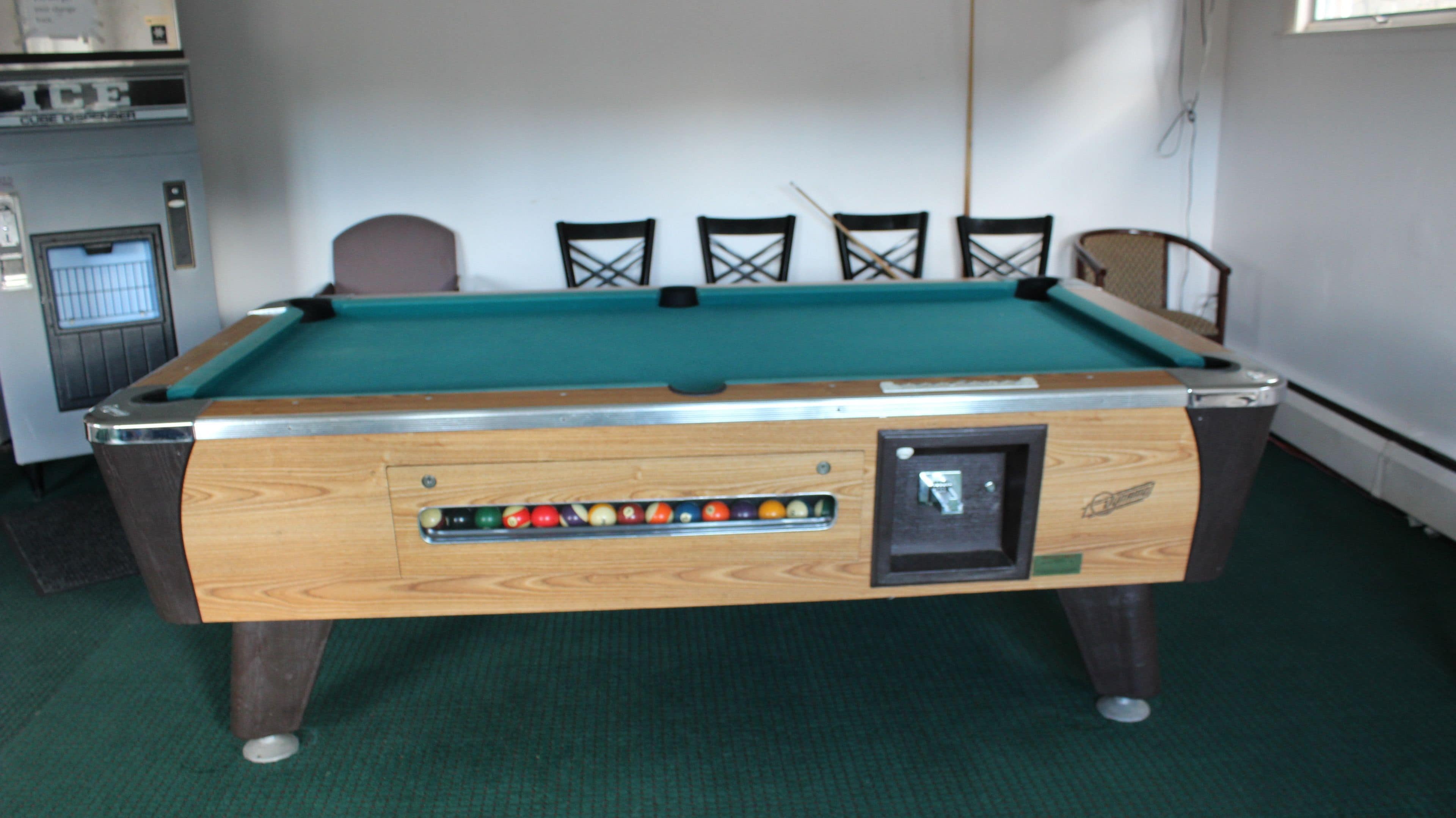 A pool table with a built-in ball rack and a cue stick leaning against the wall.