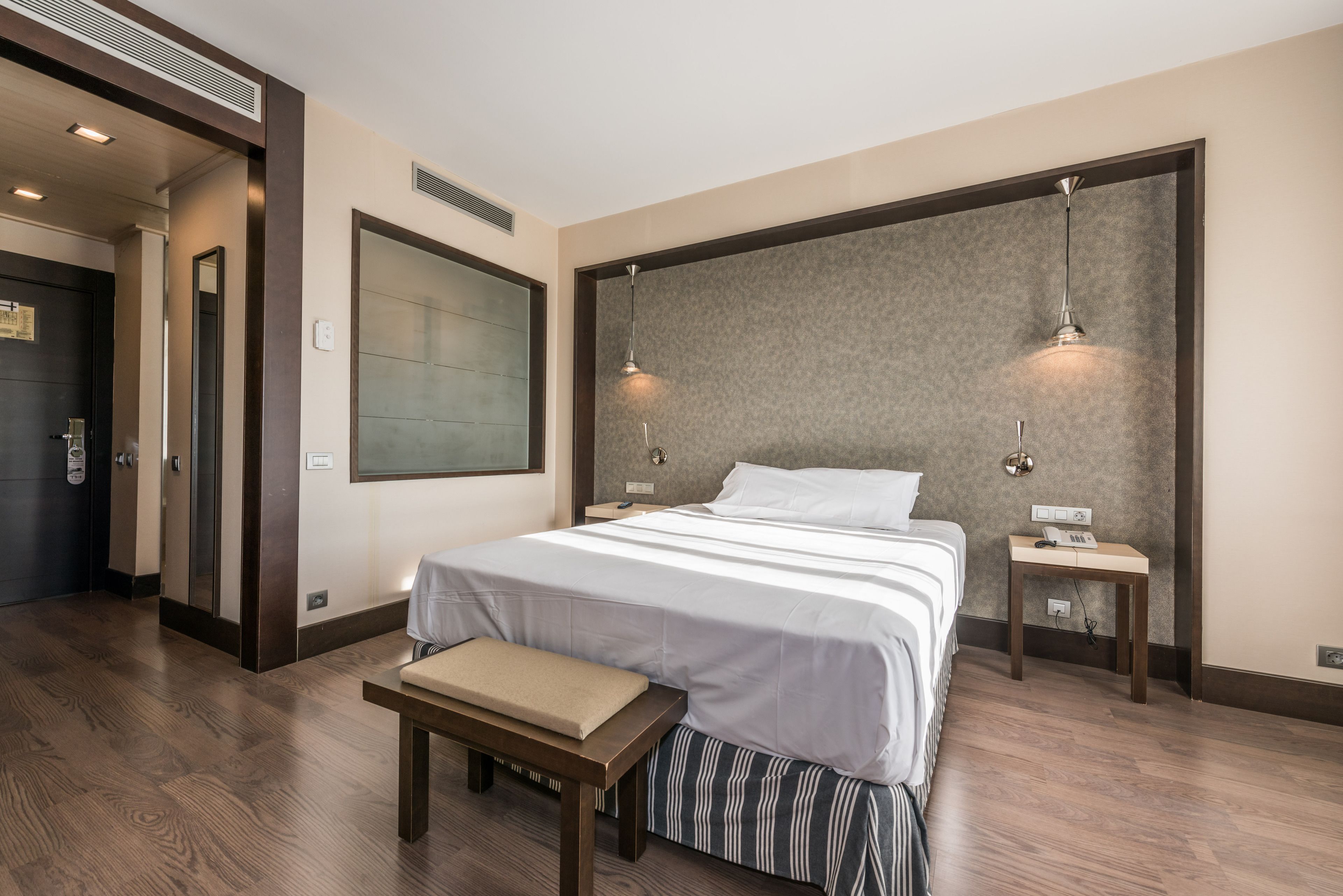 Habitación de hotel con una cama grande, un banco, dos mesitas de noche y un televisor montado en la pared.