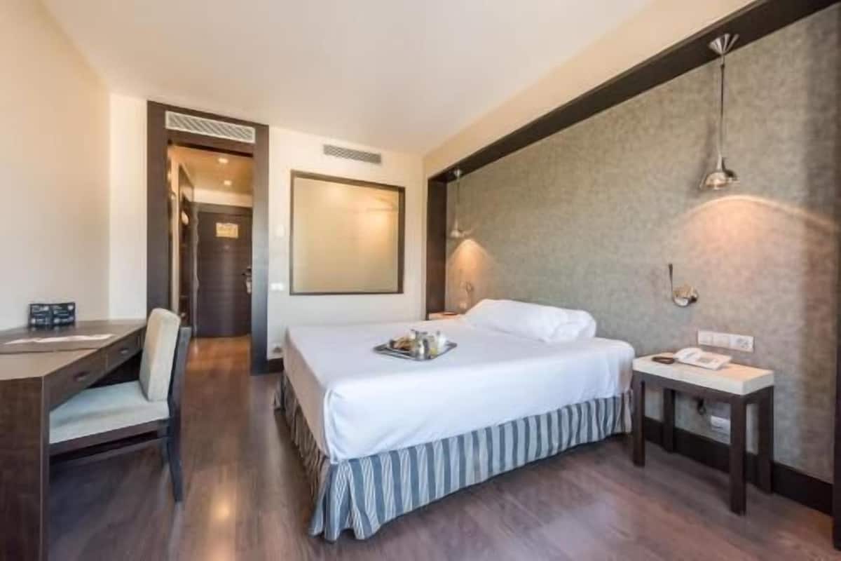 A hotel room with a bed, bedside table, desk, chair, and a large mirror.