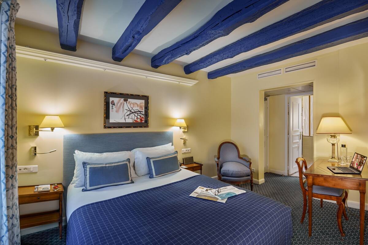 A hotel room with a large bed, two bedside tables, a chair, a desk with a laptop, and a door leading to another room.