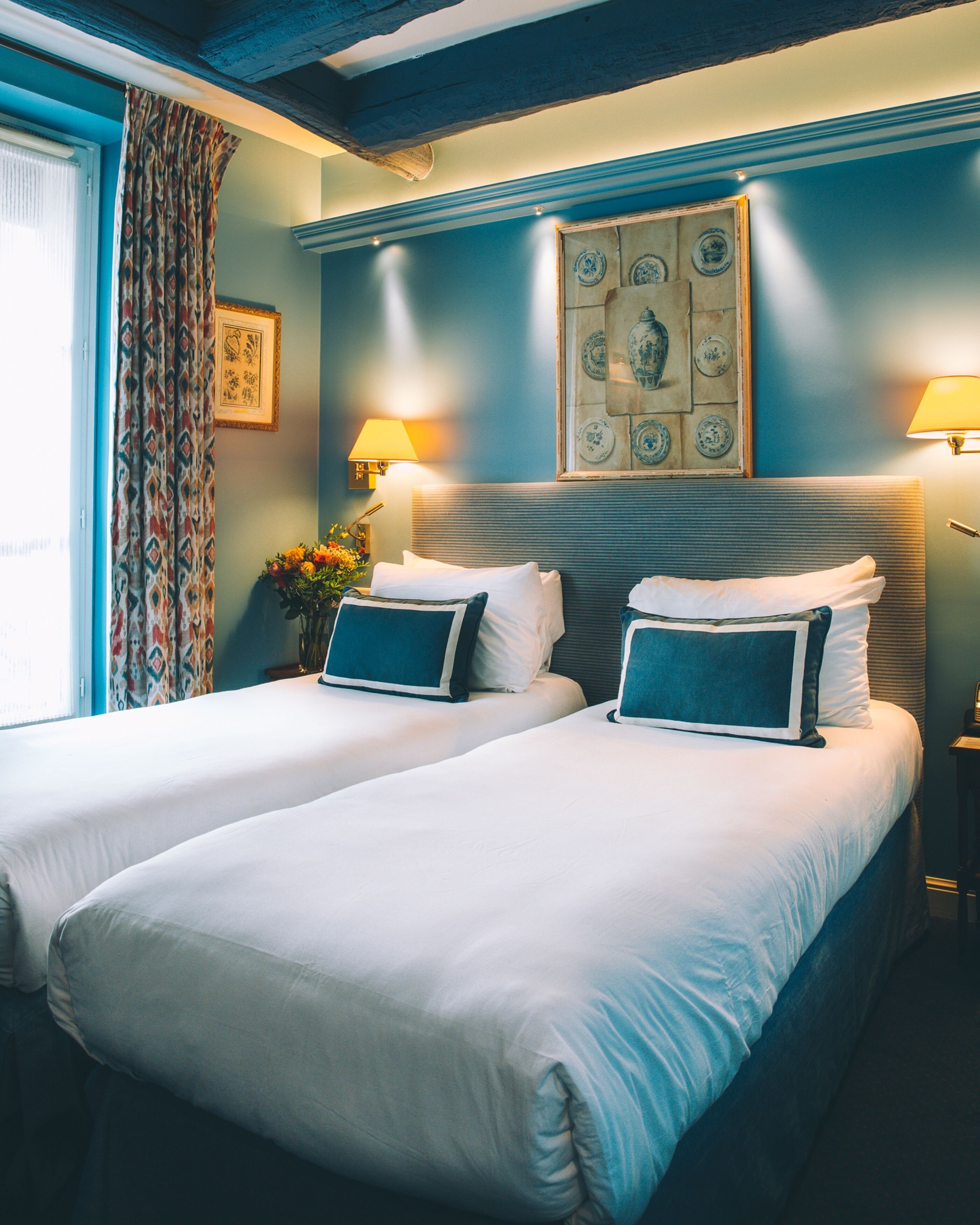 Habitación de hotel con dos camas, un ventanal con cortinas, una lámpara de pared y un plato decorativo sobre las camas.