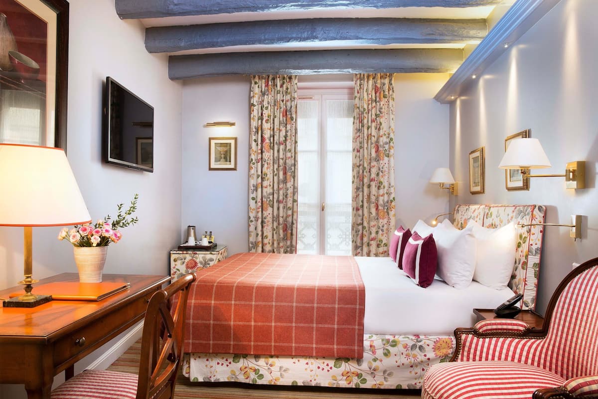 A hotel room with a bed, a desk with a lamp, a chair, a nightstand, and a TV.