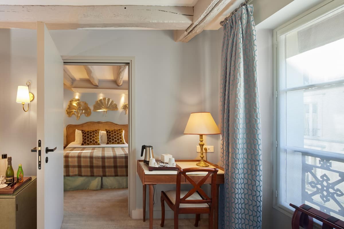 A hotel room with a bed, a desk with a lamp, a chair, a small table with bottles, and a window with curtains.