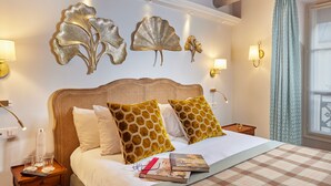 A neatly made bed with a checkered bedspread, a headboard with a woven design, and decorative wall art. There are bedside lamps, a nightstand with a bottle and glasses, and a window with curtains. - Le Relais Montmartre (Paris)