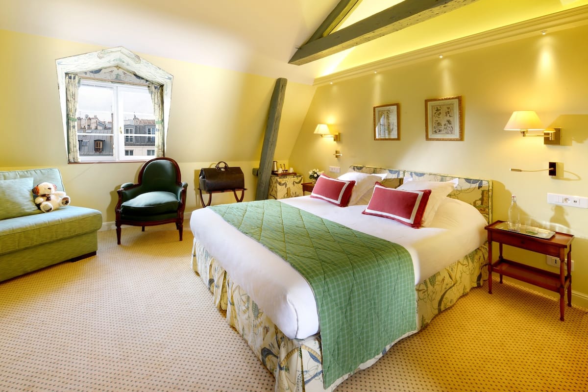 A hotel room with a large bed, a green armchair, a wooden chair, a nightstand, and a window with a view of buildings.