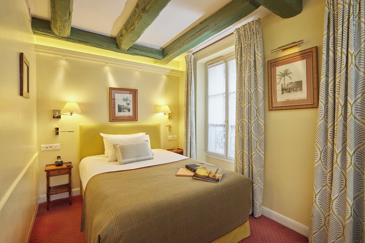 A hotel room with a bed, bedside lamps, a nightstand, a window with patterned curtains, and a framed picture on the wall.