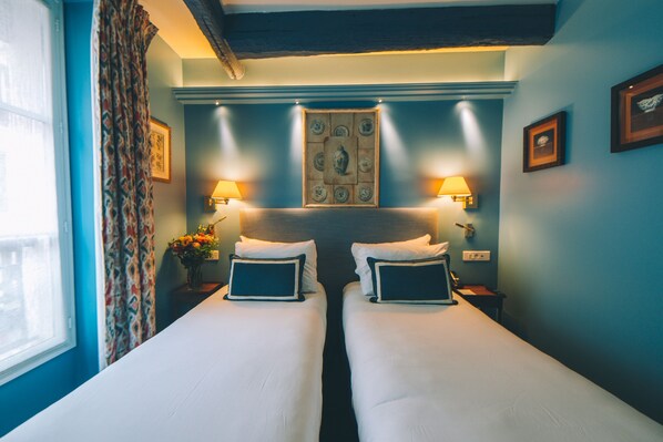A hotel room with two beds, a window with curtains, a wall-mounted lamp, and a framed picture. - Le Relais Montmartre (Paris)