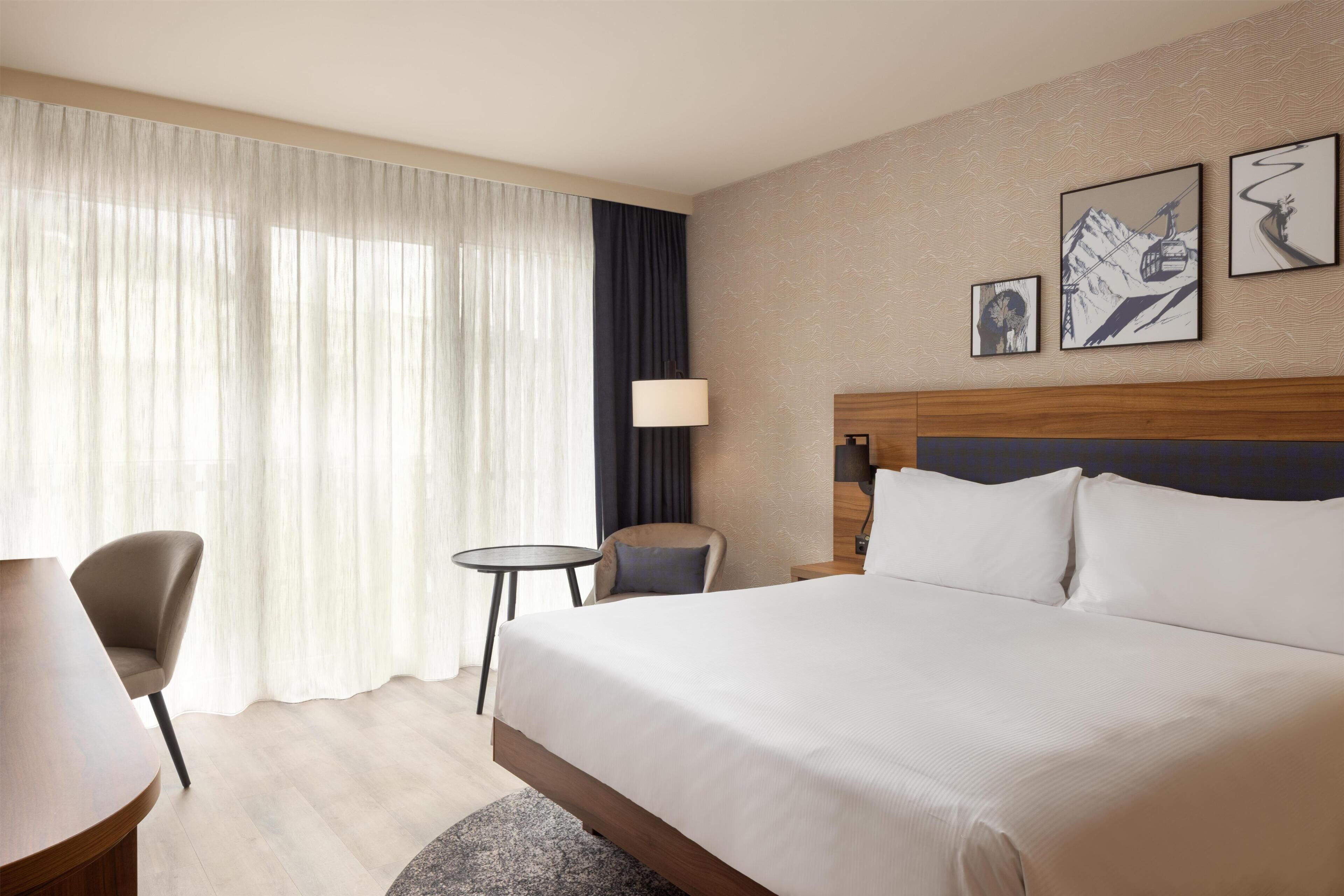 Une chambre d’hôtel avec un grand lit, un bureau, une chaise, une petite table et une fenêtre avec des rideaux. Trois tableaux encadrés sont accrochés au mur.
