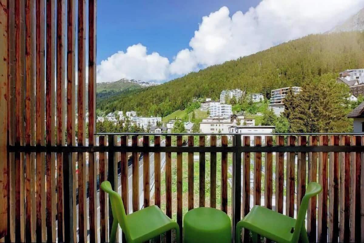 A balcony with wooden railings and two green chairs, overlooking a mountainous town with buildings and greenery.