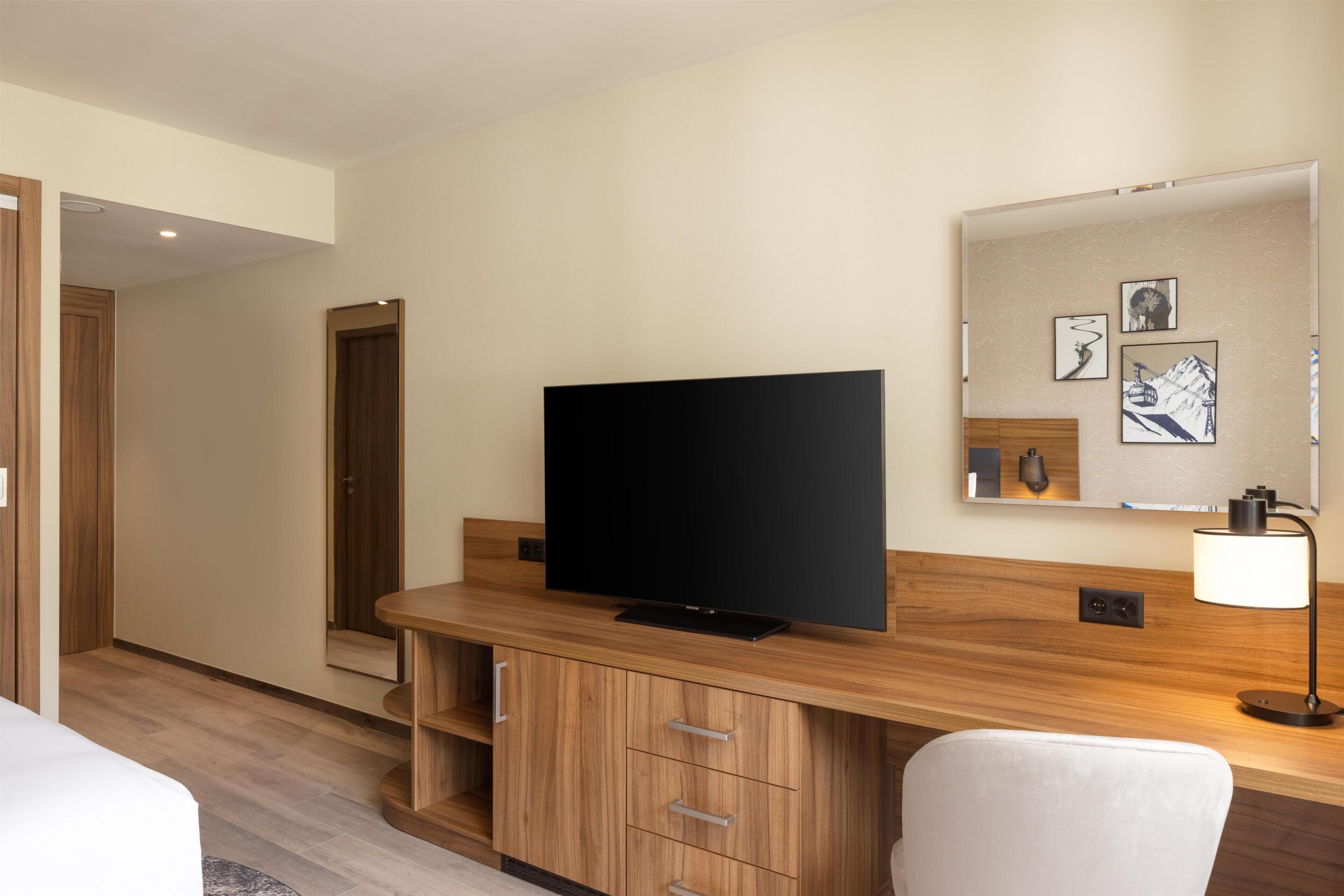 Ein Hotelzimmer mit Flachbildfernseher, Holzmöbeln, einem Spiegel mit Kunstwerk und einer Lampe.