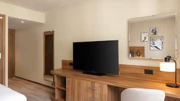 A hotel room with a flat-screen TV, wooden furniture, a mirror with artwork, and a lamp.