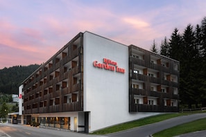 Un bâtiment moderne à plusieurs étages, sur lequel on peut voir une enseigne bien visible indiquant « Hilton Garden Inn », et qui se distingue par son architecture épurée, combinant des éléments en bois et des façades blanches.