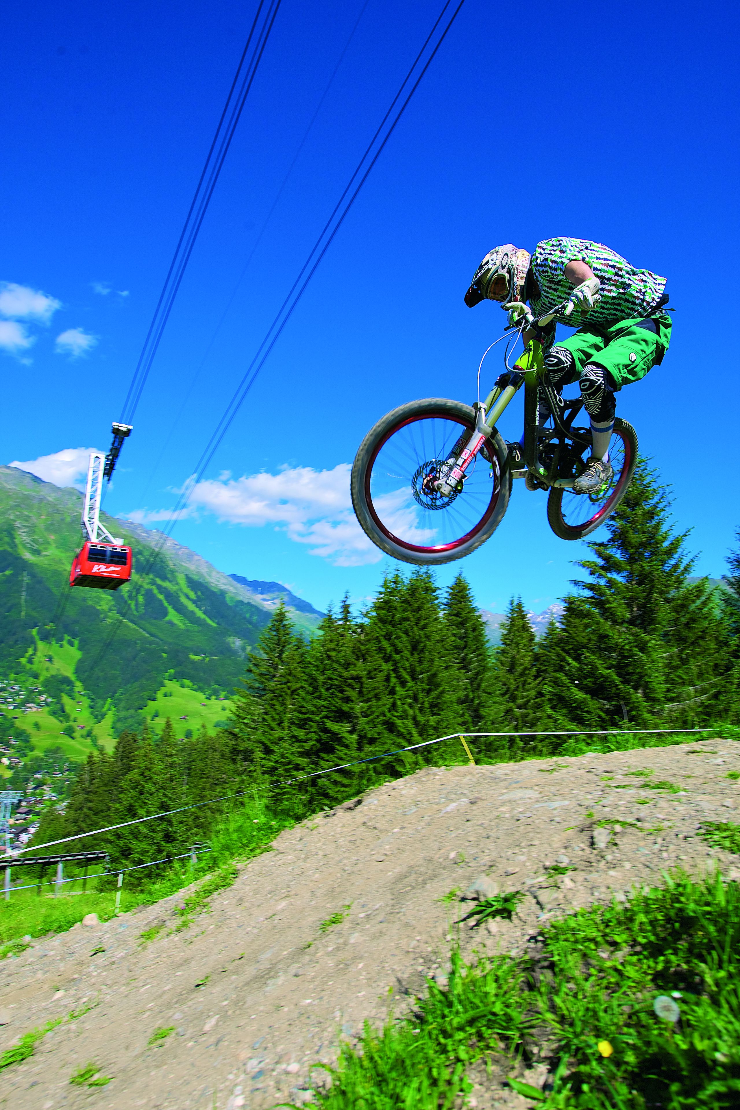 A mountain biker performing a jump with a cable car in the background.