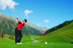 Un campo de golf con montañas al fondo, un sendero sinuoso y un golfista en pleno swing.