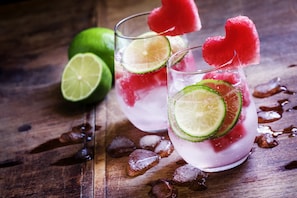 Dos vasos de una bebida rosada con rodajas de lima y una decoración en forma de corazón de sandía, cubitos de hielo y media lima sobre una superficie de madera.