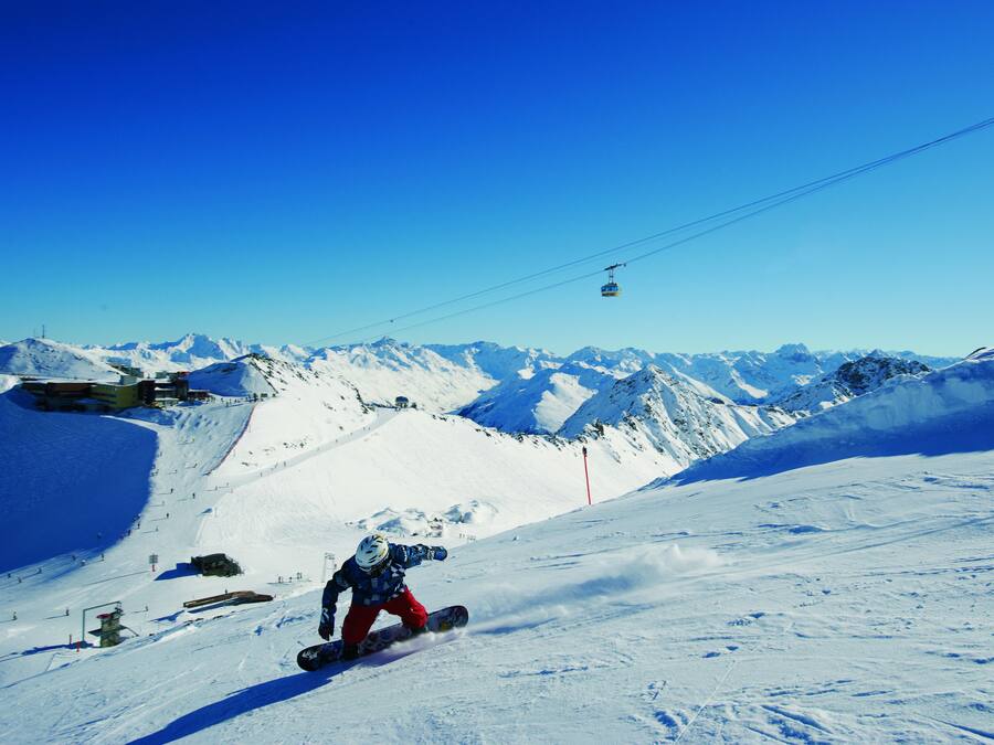 Un snowboarder en una ladera nevada con un telesilla al fondo.