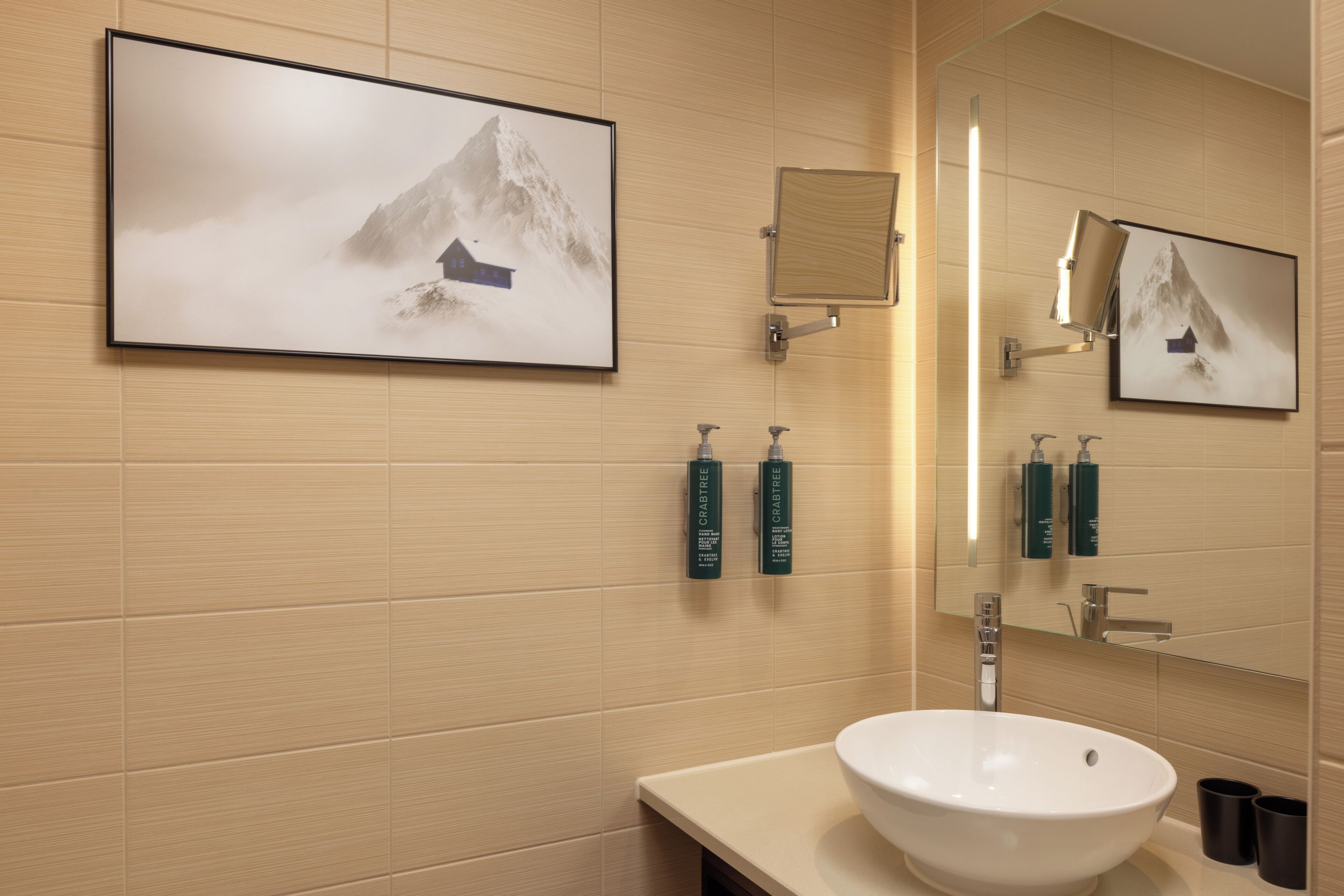 Een badkamer met een grote spiegel, een wastafel, prenten van bergen in lijsten en toiletartikelen aan de muur.