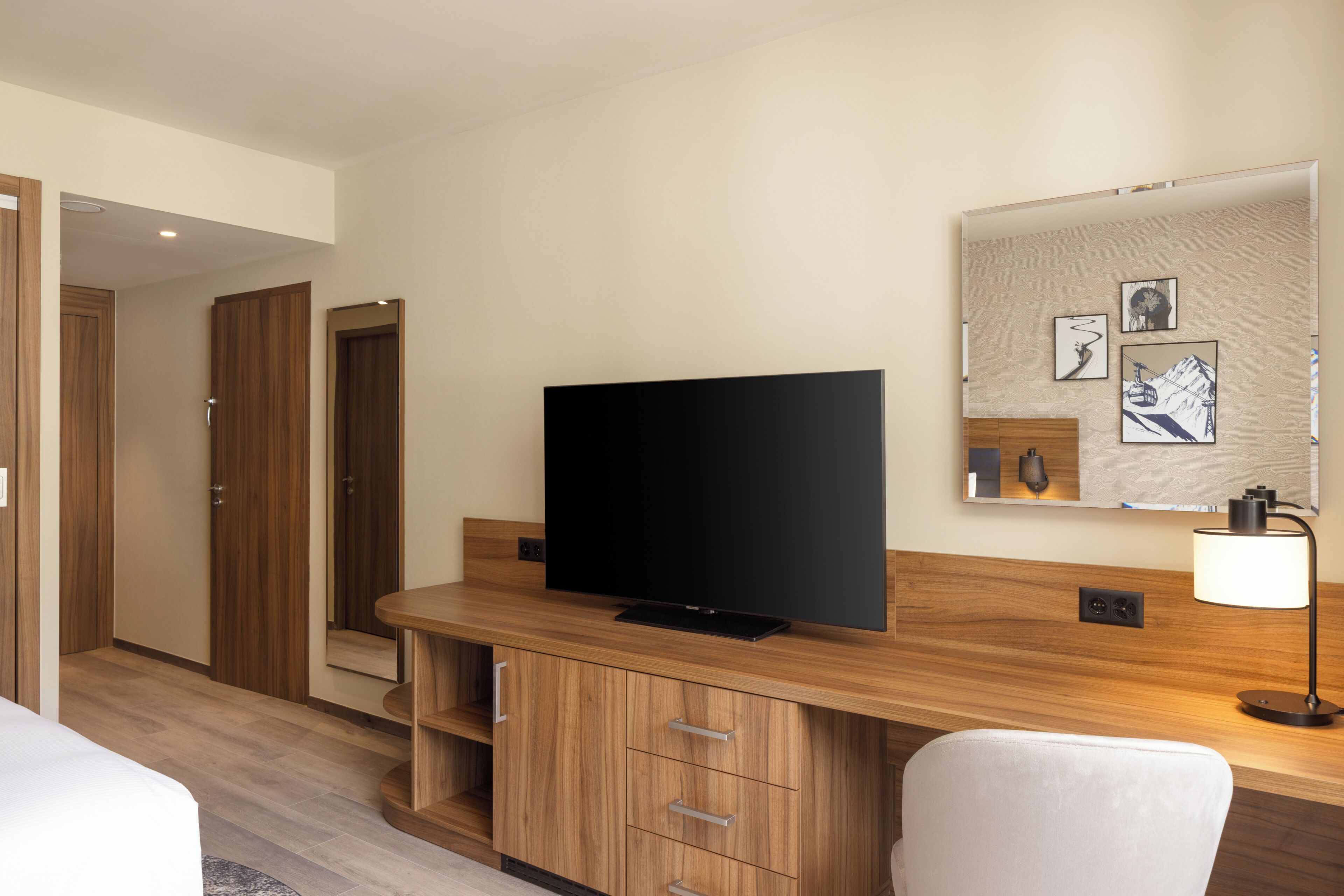 Habitación de hotel con televisión de pantalla plana, muebles de madera, un escritorio y una lámpara fijada a la pared.