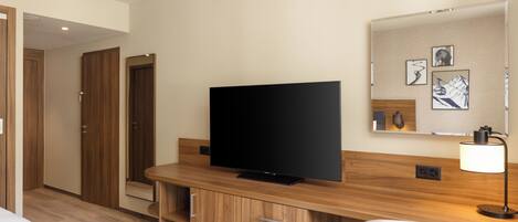 Habitación de hotel con televisión de pantalla plana, muebles de madera, un escritorio y una lámpara fijada a la pared.