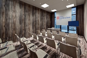 Un salón de conferencias con filas de sillas, una pantalla grande y vistas de la ciudad a través de amplios ventanales.