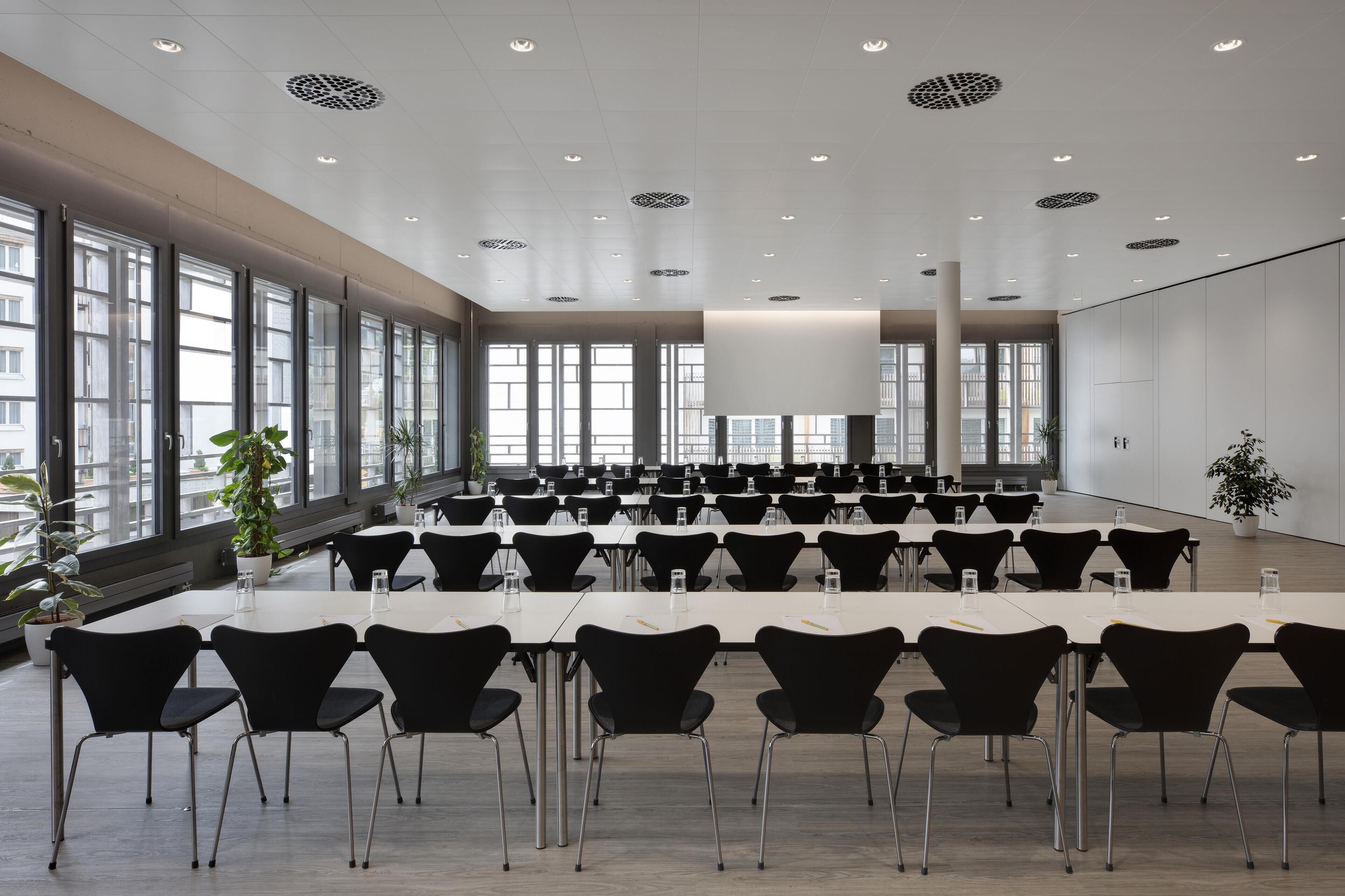 A conference room with rows of tables and chairs, large windows, and a high ceiling with recessed lighting.