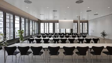 Un salón de conferencias con filas de mesas y sillas, ventanas amplias y un techo alto con iluminación empotrada.