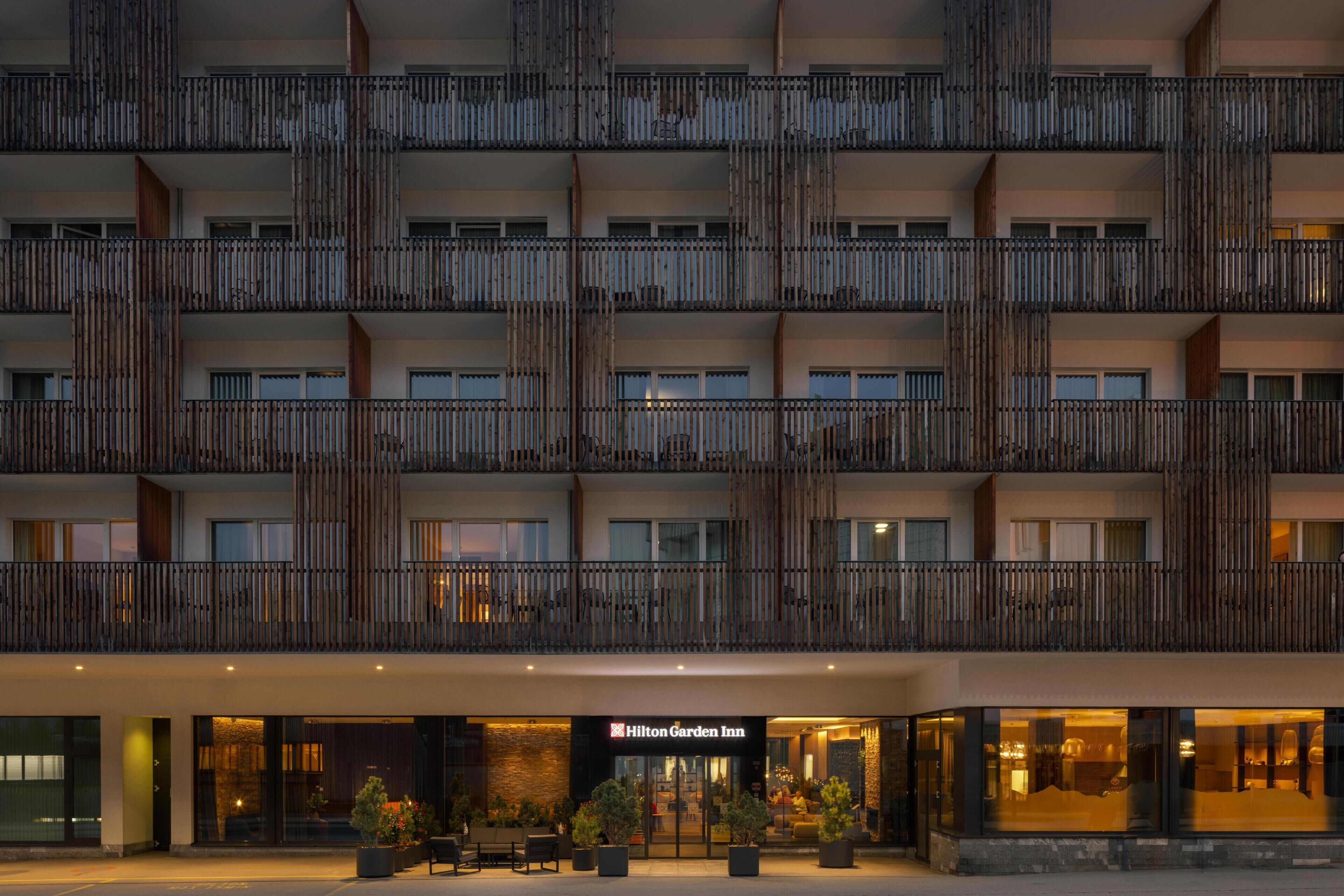 Un hotel de varios pisos con diseño moderno, que cuenta con balcones con listones verticales de madera, una entrada bien iluminada y señalización visible para Hilton Garden Inn.